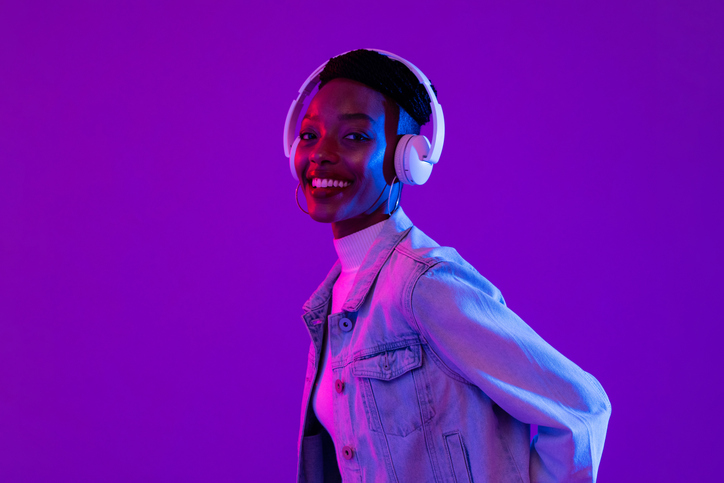 stylish happy smiling african american woman wearing headphones and listening to music in modern purple studio background